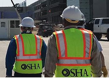Two people walking away that have hard hats and safety vests with the OSHA logo on the back