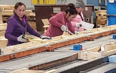 Three women wearing safety glasses and gloves working on a truss assembly line