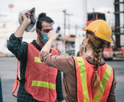 A female construction worker uses a thermometer to scan a coworkers forehead to check their temperature